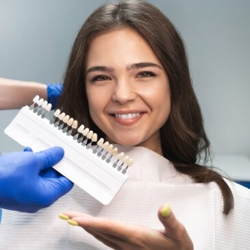 Young woman in dental chair gesturing to a row of veneers that dentist is holding