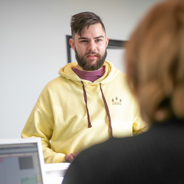 Bearded man in yellow hoodie talking to a person at a desk