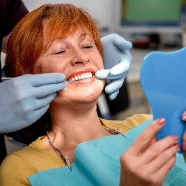 Older woman in dental chair seeing her smile in a blue hand mirror