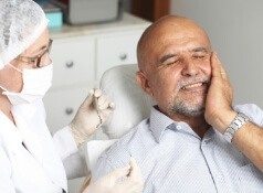 Older man in dental chair holding the side of his face in pain